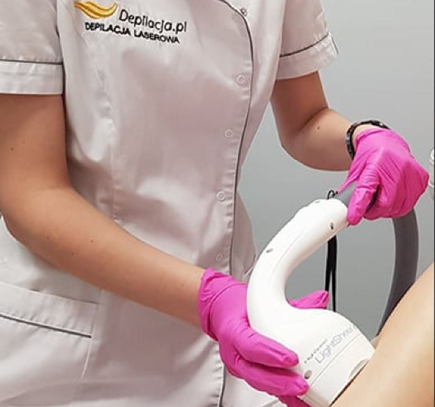 Eine Frau in einem weißen Hemd und pinken Handschuhen bedient eine weiße Laser-Haarentfernungsvorrichtung auf ihrer Haut, vor einer Wand stehend.