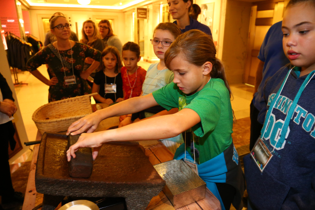 Eine Gruppe junger Mädchen steht um einen Tisch in einem Museum und untersucht verschiedene Gegenstände. Im Hintergrund hängen Kleidungsstücke an Bügeln, an der Decke befinden sich Lampen und an der Wand Rahmen.
