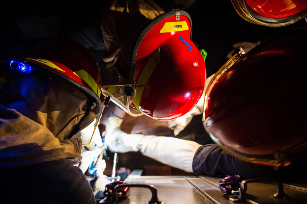 Feuerwehrleute in Helmen und Handschuhen arbeiten an einem Feuerwehrauto in einem schwach beleuchteten Bereich, wobei ein Tisch mit Vorräten rechts zu sehen ist.