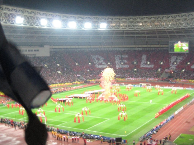 Ein Stadion mit Flutlicht, Eisengittern, Zuschauern, stehenden Personen, Werbetafeln, Anzeigetafeln, einer Sicherheitskamera, Fahnen und Deko-Elementen.