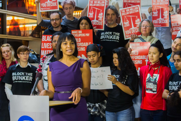 Eine Black-Lives-Matter-Aktivistin spricht in ein Mikrofon auf einem Podium, während sie eine Menge anspricht, die Schilder hält, mit Scheinwerfern und Equipment im Hintergrund.