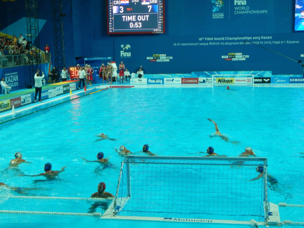 Gruppe von Menschen, die Wasserball in einem Pool während der 2015 FINA Weltmeisterschaft in Kazan spielen, mit Zuschauern und Eventbeschilderung im Hintergrund.