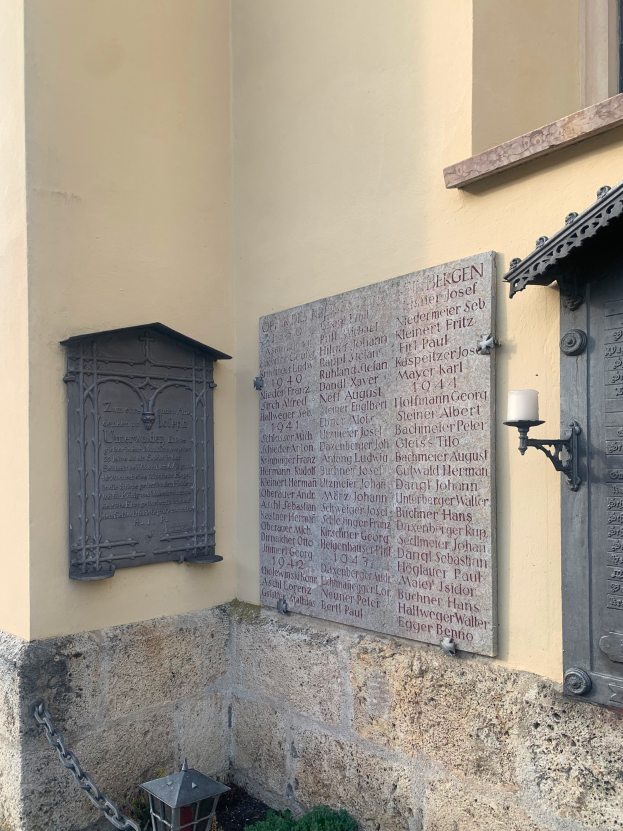 Ein Gebäude mit einer Holocaust-Gedenktafel, einem Fenster, einer Tür, einer Außenbeleuchtung, einer Kette und umgebenden Pflanzen.
