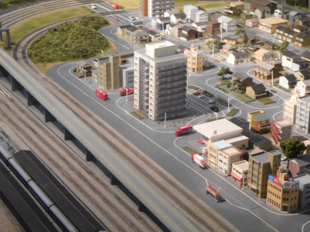 Ein Miniaturmodell einer Stadt mit einem Zug auf Schienen, umgeben von Gebäuden, Bäumen, Straßen, Fahrzeugen, Strommasten, Gras und einem Wolkenkratzer.