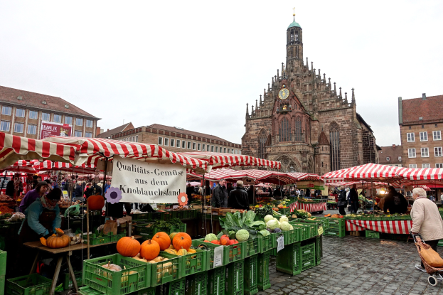 Ein belebter Markt in Nürnberg, Deutschland, mit verschiedenen Früchten und Gemüse, Menschen mit Taschen, Zelten, Gebäuden mit Fenstern, einem Uhrenturm und dem Himmel im Hintergrund.