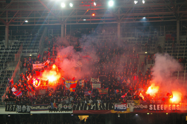 Eine große Menschenmenge in einem Stadion hält Fahnen und Banner, mit Leuchtraketen, die Rauch erzeugen, unter einer Decke mit Deckenleuchten und Metallrahmen.