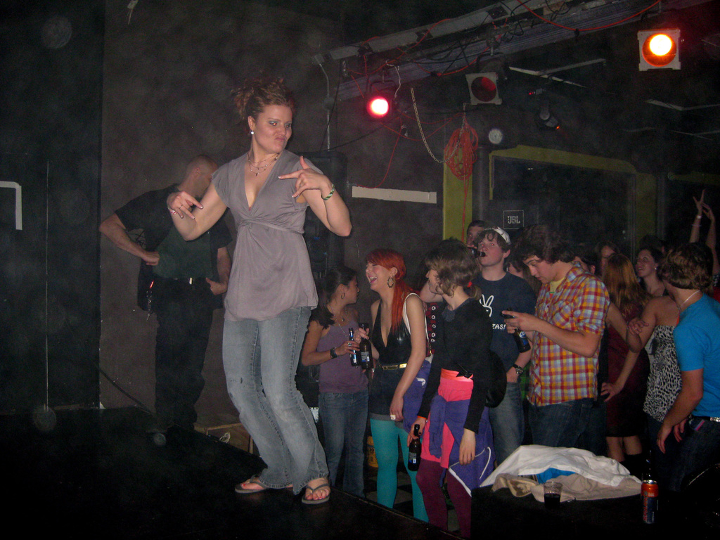 Eine Gruppe von Menschen auf einer sozialen Veranstaltung, wobei einige Bierflaschen halten, eine Person tanzt auf der Bühne und ein Mann in einer Mütze trinkt aus einer Flasche.