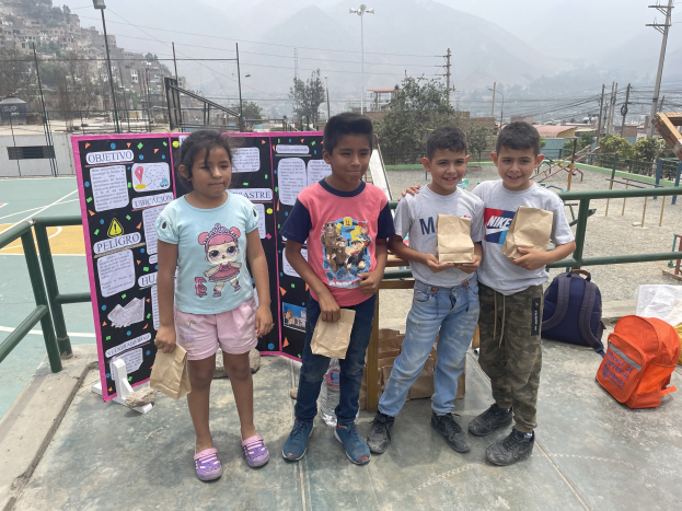 Gruppe von Kindern, die Papiertüten auf einem Basketballfeld halten, mit Tüten und Gegenständen auf dem Boden, einer Tafel mit Text links und einem Zaun, Pfosten und Gebäuden im Hintergrund.