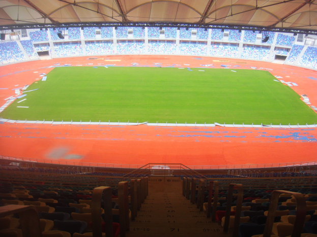Ein großes Stadion voller Zuschauer mit Sitzreihen, Geländern, Treppen und einem Dach, mit dem Spielfeld in der Mitte sichtbar.