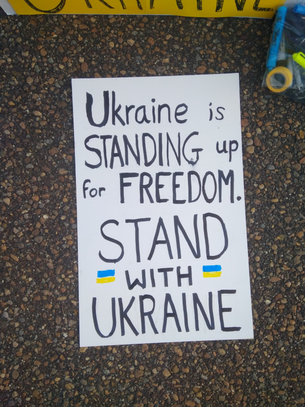 Weißer Zettel mit dem Text "Ukraine steht für Freiheit, steht mit Ukraine" auf dem Boden vor einer gelben Fahne, mit einer Plastikhülle mit verschiedenen Gegenständen rechts daneben.