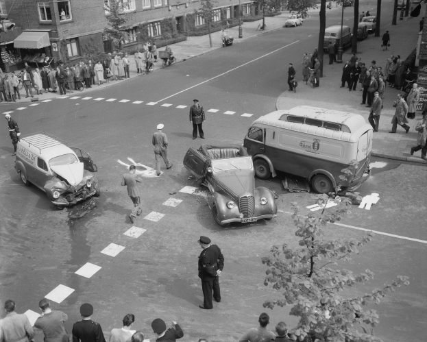 Schwarze und weiße Szenerie eines Autounfalls an einer Straßenkreuzung mit mehreren Fahrzeugen, darunter ein Van, und einer Gruppe von Menschen, die danebenstehen, mit Gebäuden, Bäumen, Pfählen und Texttafeln im Hintergrund.