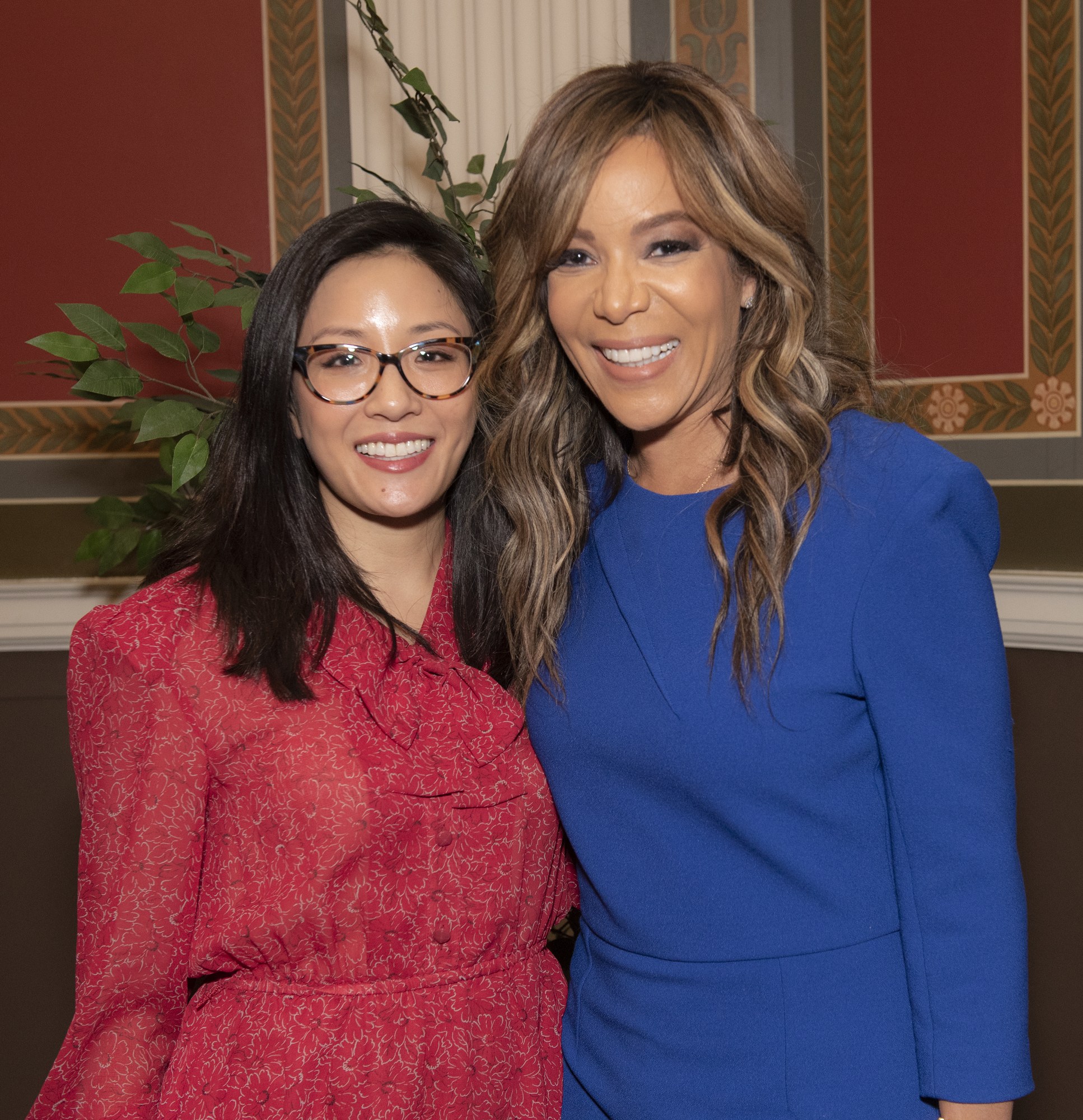 Zwei Frauen, die zusammen auf der 2019 Women in Film Pre-Oscar-Luncheon lachen, eine in einem blauen Kleid und die andere in einem roten Kleid mit Brille, stehen neben einer Pflanze und einem Vorhang.