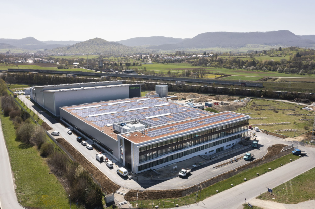 Luftaufnahme eines großen Industriegebäudes mit Solarpaneelen auf dem Dach, umgeben von Fahrzeugen, Bäumen, Gras, Strommasten und Stromleitungen, vor Bergen und einem klaren blauen Himmel.