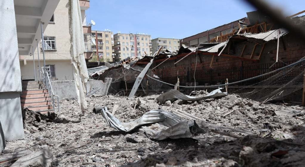 Die Nachwirkungen eines israelischen Luftangriffs in Gaza-Stadt zeigen ein eingestürztes Gebäude, das zu Schutt und Asche geworden ist, in einer Straße mit sichtbaren Trümmern, Metallstangen, einem Zaun, einer Treppe, benachbarten Gebäuden, Bäumen und einem bewölkten Himmel.
