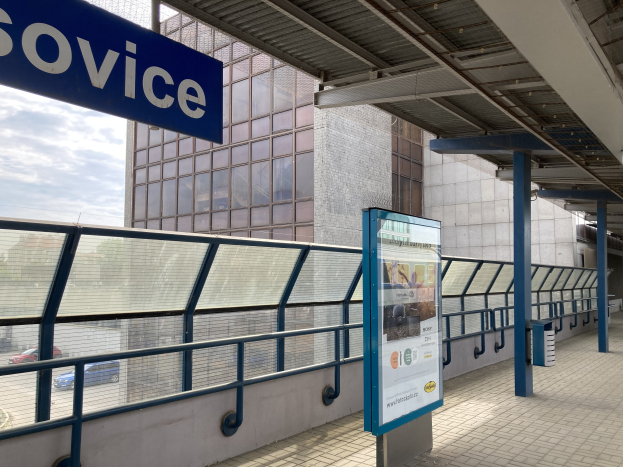 Haltestelle mit einem "Service"-Schild, Infotafel, Metallzaun, Pfosten, Dach mit Deckenleuchten, nahegelegene Gebäude, Fahrzeuge auf der Straße, Bäume und ein bewölkter Himmel.