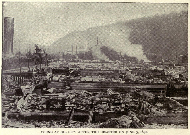 Schwarzes und weißes Foto einer Stadt nach einer Katastrophe am 5. Juni 1892, das Gebäude, Pfosten, Rauch und einen Hügel im Hintergrund zeigt, mit Text am unteren Rand.