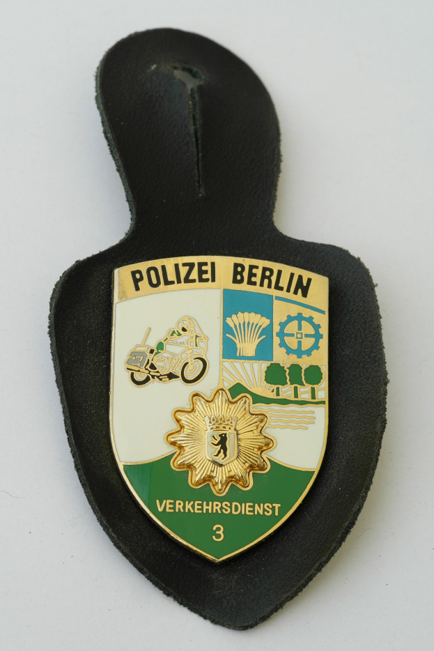 Ein schildförmiges Polizeibadge mit der Aufschrift 'Polizei Berlin' auf blauem Hintergrund, weißem Rand, zentralem weißen Stern und schwarzer fetter Schrift, das auf einer Oberfläche ruht.