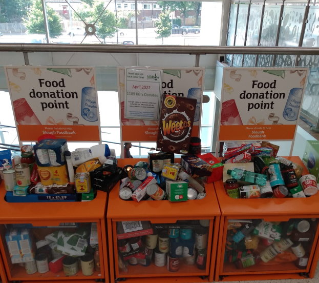 Orangene Schränke mit Lebensmittelspenden gefüllt, darunter Flaschen, Dosen und Kartons, mit Informationsschildern und einem Blick auf Fahrzeuge, Bäume, Gebäude und Himmel durch Glasfenster.