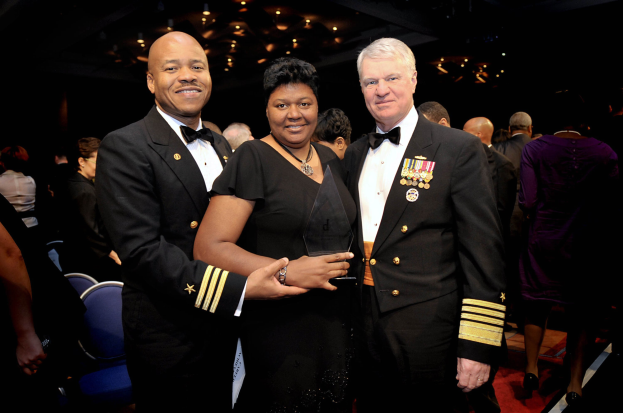 Zwei Männer in schwarzen Blazern und weißen Hemden stehen neben einer Frau in einem schwarzen Kleid, die eine Trophäe hält, auf einem roten Teppich, während andere sitzen und stehen und der Hintergrund von beleuchteter Decke erhellt wird.