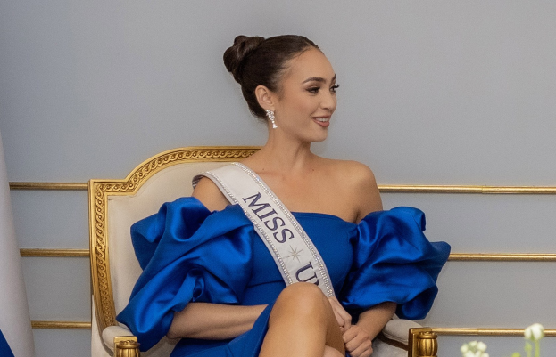 Frau in einem blauen Kleid mit einem "Miss Universe 2019"-Band, die auf einem Stuhl sitzend und strahlend von Blumen und einer Wand im Hintergrund umgeben ist.