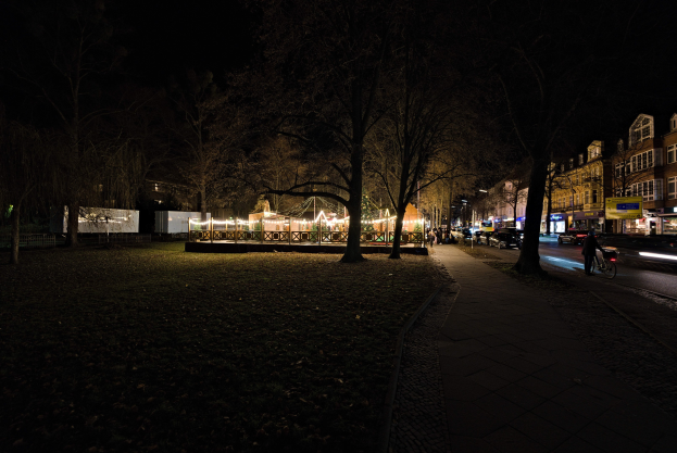 Eine Straße in einer Stadt bei Nacht mit fahrenden Autos, Laternen, Bäumen, Gebäuden mit erleuchteten Fenstern, einem Radfahrer im Vordergrund und einem dunklen Himmel.