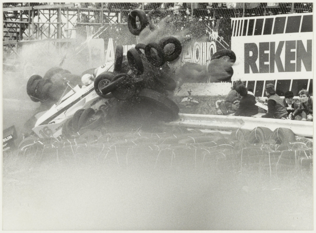 Schwarz-weißes Foto eines verunglückten Rennwagens umgeben von Reifen, mit Menschen rechts sitzend und einer Tafel mit Text im Hintergrund.