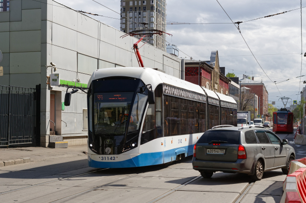 Blauer und weißer Straßenbahnwagen fährt auf einer Stadtstraße mit hohen Gebäuden, Autos, Bäumen, Strommasten und Drähten im Hintergrund unter einem bewölkten Himmel.