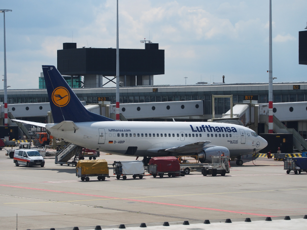 Lufthansa Airbus A320-200 Flugzeug auf dem Frankfurter Flughafen geparkt mit Bodenfahrzeugen und Wagen vorne, ein Gebäude und Laternenmasten im Hintergrund unter einem klaren blauen Himmel.