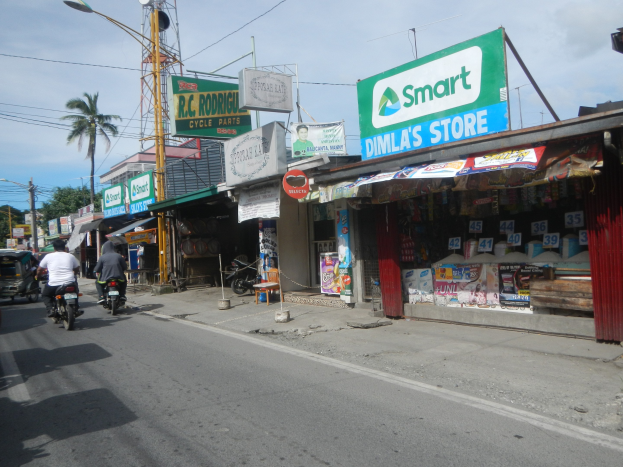 Eine belebte Straße mit Motorradfahrern, Autos, Gebäuden, Straßenschildern, Strommasten, Kabeln, Straßenlaternen, Bäumen und einer bewölkten Himmel.