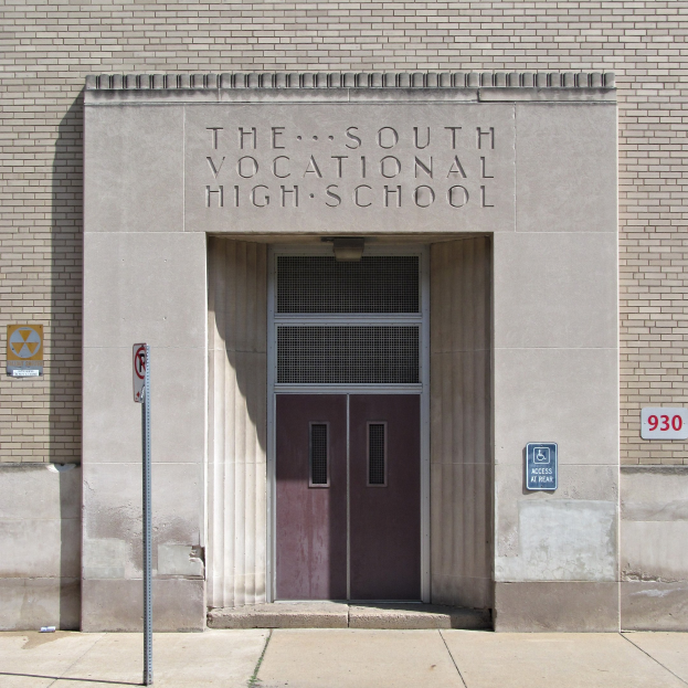 Außenansicht der Südlichen Berufsschule mit einer Tür, Schildern an der Wand und einem Weg, der zur Eingangstür führt.