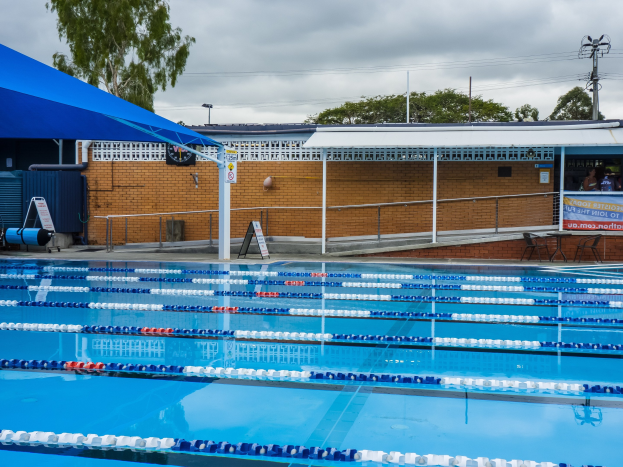Ein großes Schwimmbad mit Bahnentrennung, umgeben von einem Zaun, einer Hütte, einem Banner, Stühlen, einem Tisch, einem Schild, Pfählen, einer Straßenlaterne, Drähten, Bäumen und einem bewölkten Himmel.