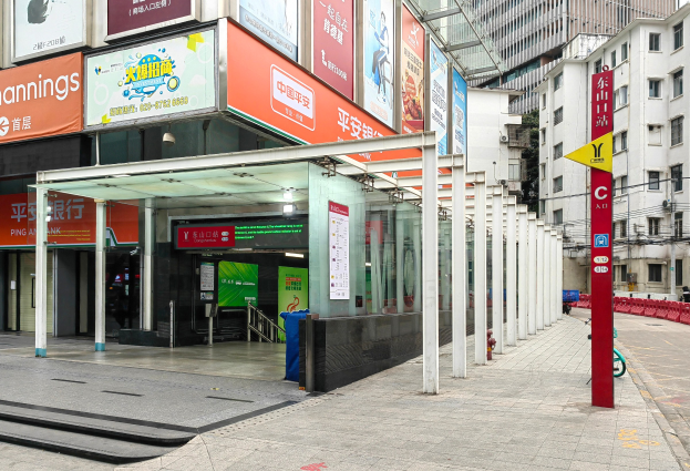Eine Stadtstraße mit einer Bushaltestelle, Gebäude auf beiden Seiten, eine Straße mit einem Müllcontainer, rote und gelbe Schilder mit Text, Fußgänger auf dem Gehweg und eine Rolltreppe auf der linken Seite.