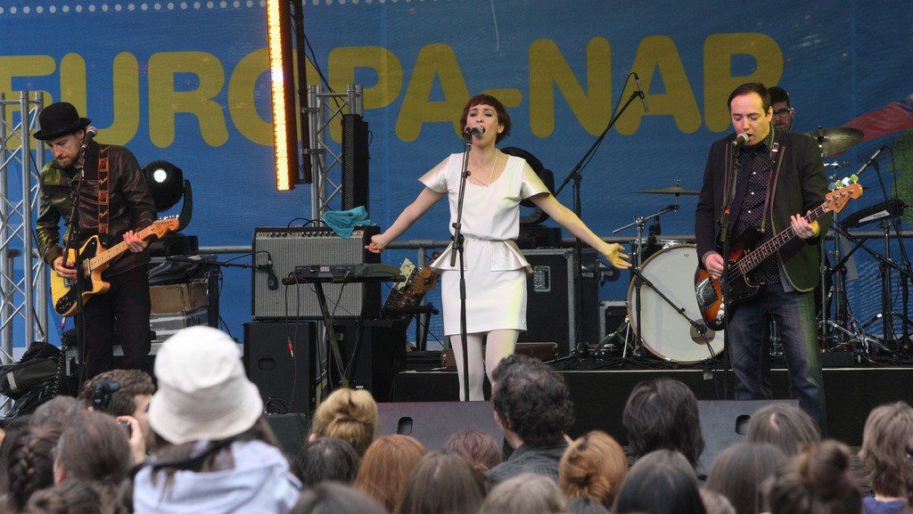 Vier Personen auf der Bühne: eine Frau in einem weißen Kleid, die singt, zwei Männer, die Gitarren spielen, und eine Menge darunter, mit einer Wand und Lautsprechern im Hintergrund.