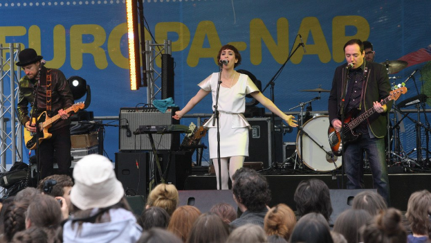 Vier Personen auf der Bühne: eine Frau in einem weißen Kleid, die singt, zwei Männer, die Gitarren spielen, und eine Menge darunter, mit einer Wand und Lautsprechern im Hintergrund.