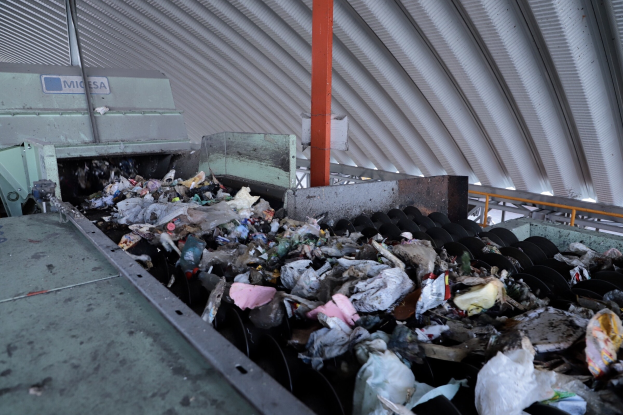 Großer Haufen Müll auf einem Förderband in einer Recyclinganlage mit Maschinen und einem Metalldach.