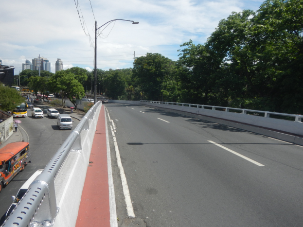 Autobahn mit fahrenden Autos und Bussen, gesäumt von Geländern, Laternen, Bäumen, Gebäuden und einem bewölkten Himmel.