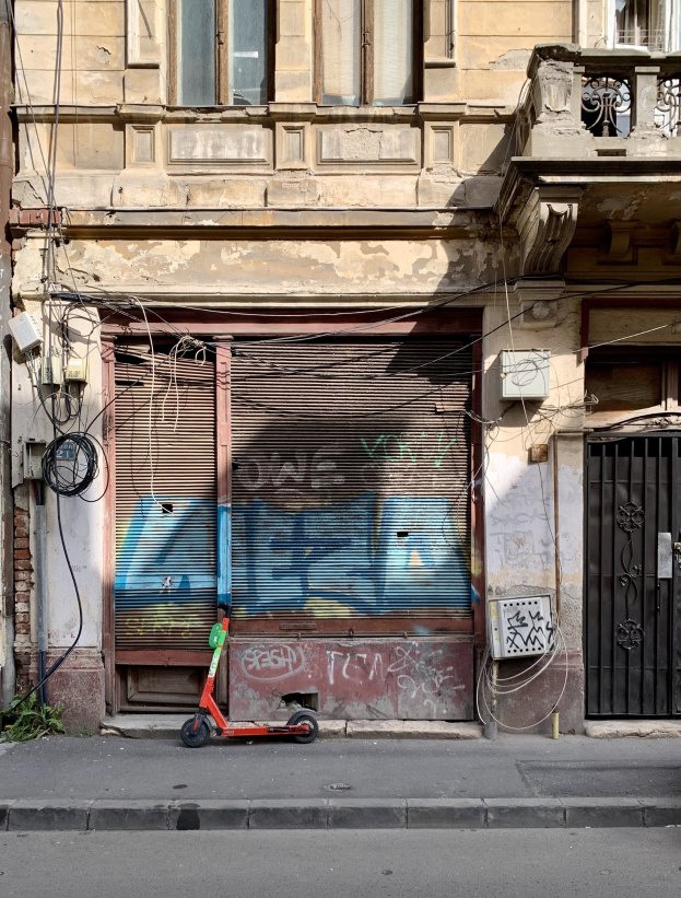 Ein roter Roller, der auf einer Straße neben einem Gebäude mit Fenstern, Rollläden und einem Tor geparkt ist, mit sichtbaren Straßenkabeln und Text unten.