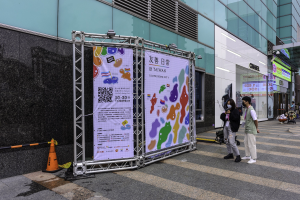 Eine Gruppe von Menschen mit Masken geht eine Straße neben einem hohen Gebäude entlang, mit Schildern auf einem Metallrahmen, Verkehrspfosten und einer Schautafel, Bäumen und einem bewölkten Himmel.