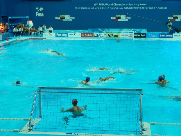Eine Gruppe von Menschen, die Wasserball in einem Pool mit einem Netz spielen, mit Zuschauern im Hintergrund und Tafeln mit Text und Logos, die die 2015 FINA Weltmeisterschaft in Kazan anzeigen.