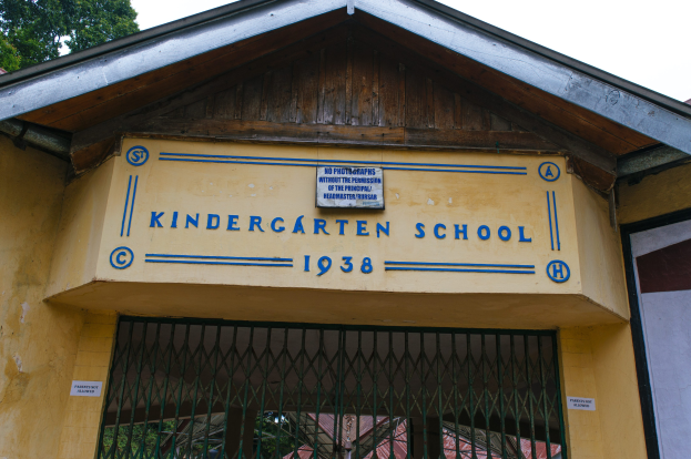 Gelbes Gebäude mit einer "Kindergarten-Schule"-Schilder und einer Wandtafel, darunter ein Gitter und Bäume vor einem klaren blauen Himmel.