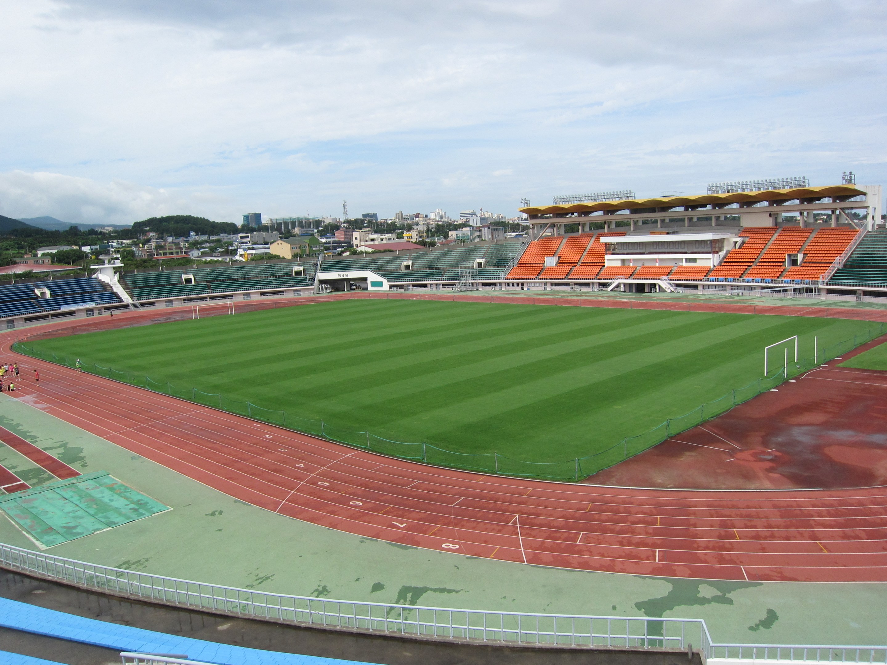Ein großes Stadion mit einem Fußballfeld, umgeben von Gebäuden, Bäumen, Hügeln und einem klaren blauen Himmel, mit ein paar Menschen auf dem saftig grünen Rasen.