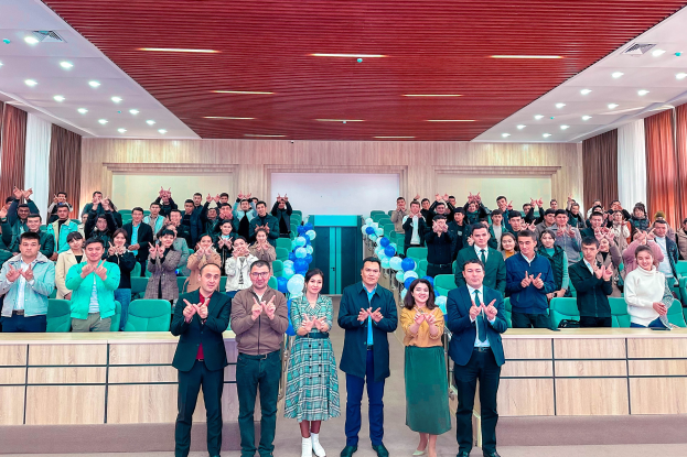 Eine Gruppe von Menschen steht vor einem Publikum, einige klatschen, in einem Raum mit Stühlen, Tischen, Luftballons, Vorhängen und Deckenleuchten, bei der Eröffnung einer neuen Schule.