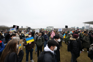 Eine große Gruppe von Menschen mit Masken und Schildern demonstriert vor einem Gebäude in Washington, D.C., mit Bäumen, Gebäuden und einem Schuppen im Hintergrund.