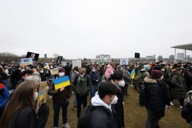 Eine große Gruppe von Menschen mit Masken und Schildern demonstriert vor einem Gebäude in Washington, D.C., mit Bäumen, Gebäuden und einem Schuppen im Hintergrund.