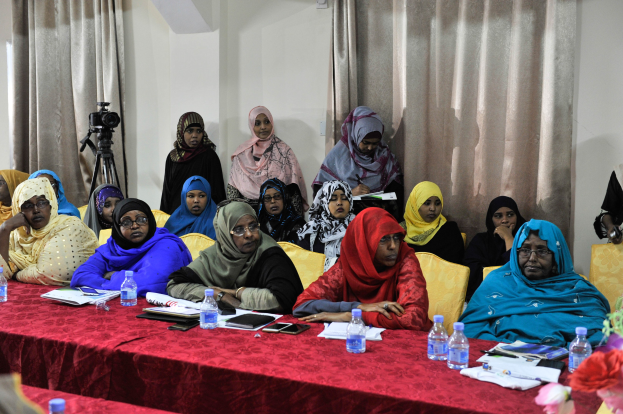 Eine Gruppe von Frauen sitzt an einem Tisch mit Flaschen, Büchern, Handys, Papieren, Stiften und einer Blumenvase und diskutiert vor einer Vorhangwand.