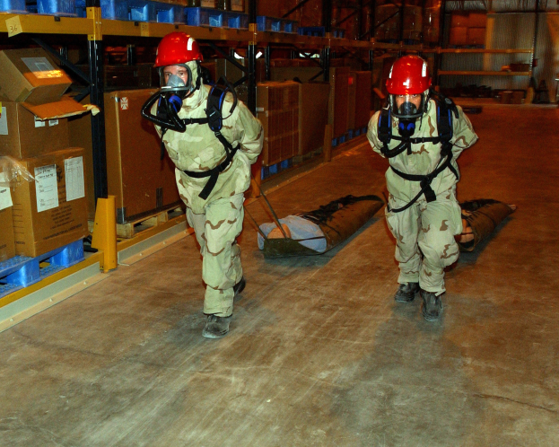 Zwei Militärangehörige in Gasmasken und roten Helmen tragen eine Trage durch ein Lagerhaus voller Kartons und Regale.