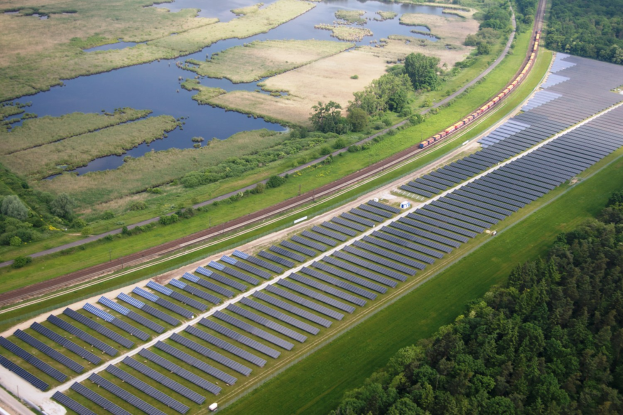 Luftaufnahme eines Solarparks mit Solarpanelen, umgeben von Bäumen, Gras, Pflanzen und Wasser, mit einem Zug auf einer nahen Bahnlinie.