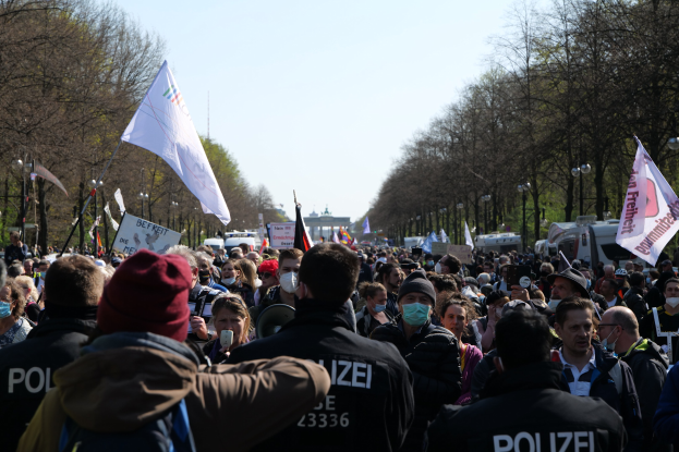 Eine große Gruppe von Menschen vor einer Reihe von Polizeibeamten bei einer öffentlichen Demonstration, mit Transparenten, Fahnen und Fahrzeugen im Hintergrund.