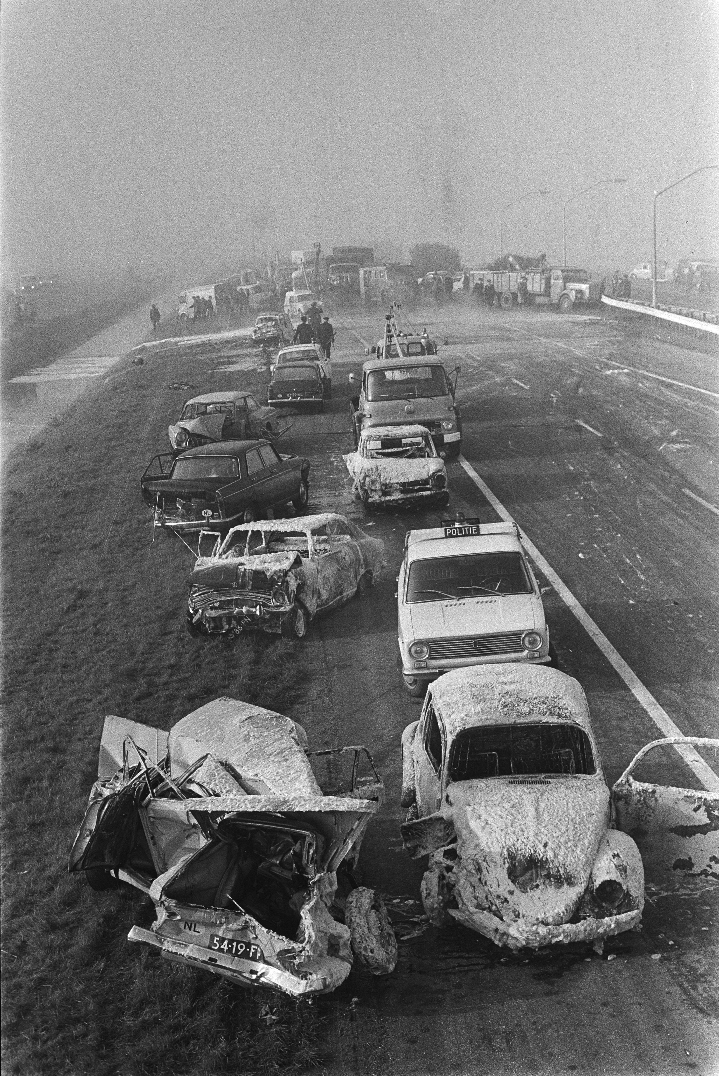Ein Schwarz-Weiß-Foto von mehreren verunglückten Autos am Straßenrand, umgeben von Gras, Laternen und einigen Menschen, mit Bäumen und einem klaren Himmel im Hintergrund.