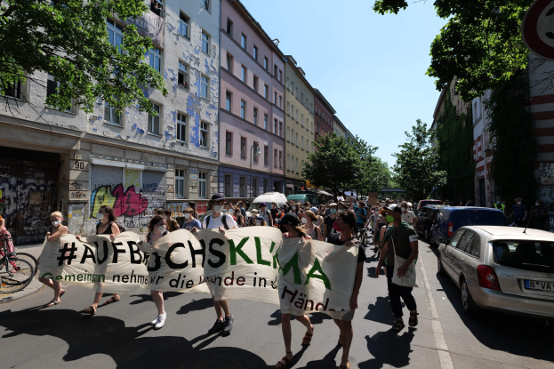 Eine Gruppe von Menschen, die auf einer Stadtstraße marschieren und ein Banner halten, mit parkenden Autos auf der rechten Seite, einem Radfahrer auf der linken Seite und Gebäuden, Bäumen und einem Schild im Hintergrund unter einem sichtbaren Himmel.